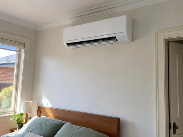 Wall-mounted split system running above a wooden bed in a bright bedroom in Kilburn